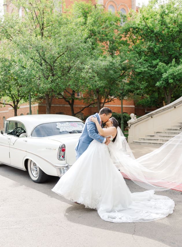 Groom dips bride with her veil flowing in front of a vintage car outside San Antonio wedding venue