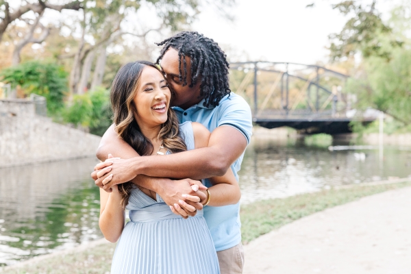 Man embraces his fiancée from behind holding on to her hands. There is a beautiful river and bridge in the background of this San Antonio engagement photo. 