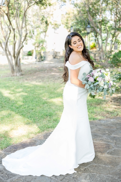 Bride closes her eyes to show work of a wedding makeup artist in San Antonio. She holds her bouquet with both hands with an off the shoulder dress with a small train. The background has beautiful green grass, cobblestone walkway, and some trees.