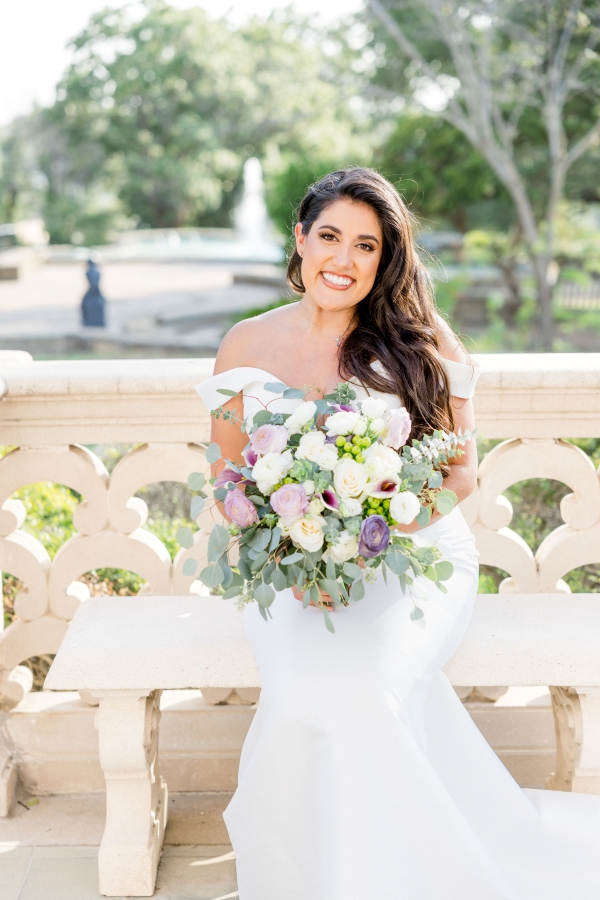 Bride in an off the shoulder sleeved dress sits on a stool in front of a grove of trees. She had a wedding makeup artist in San Antonio do professional make up for her wedding day.