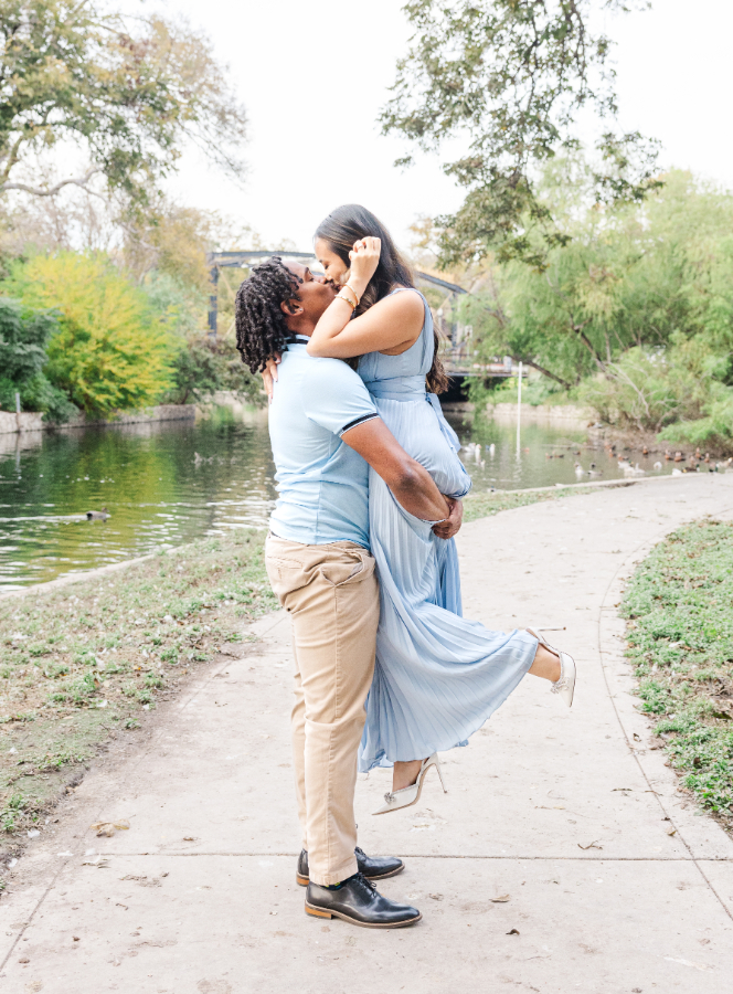 The groom to be in this San Antonio engagement photo session holds up his bride to be as she kisses him with one leg up. They're walking along a river on a paved path with a bridge in the background.