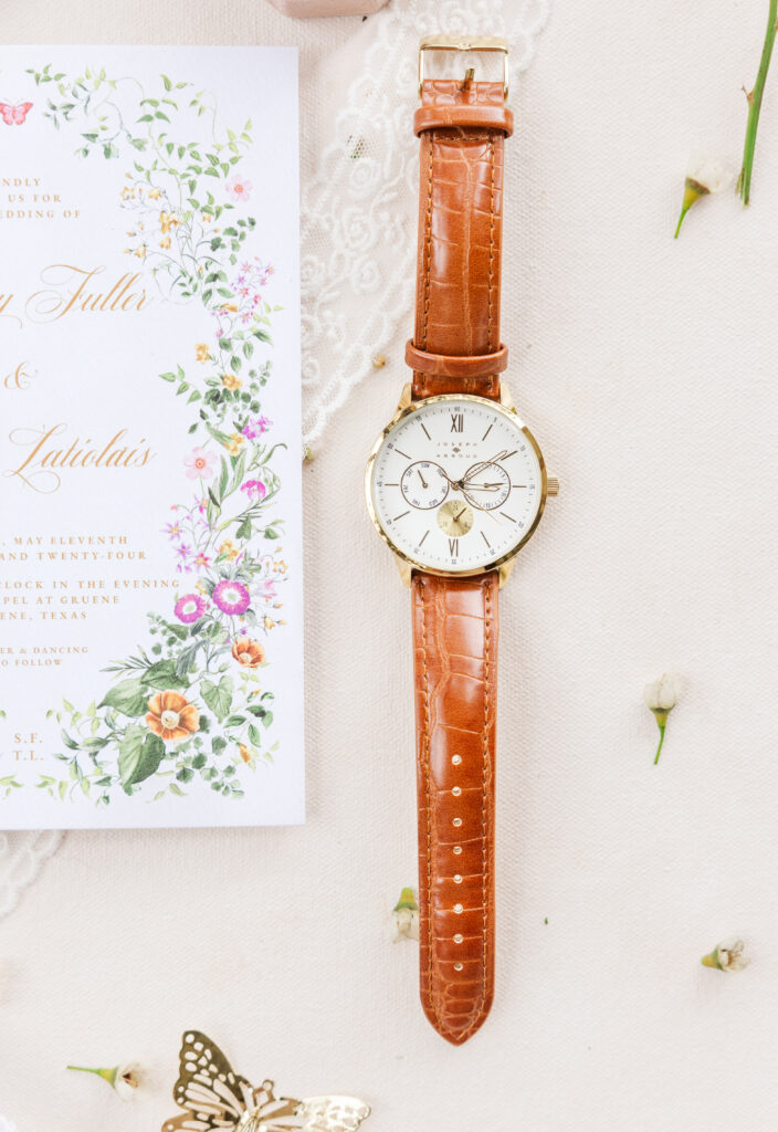 a watch sits next to an invitation with small white flowers scattered for wedding flat lay photo