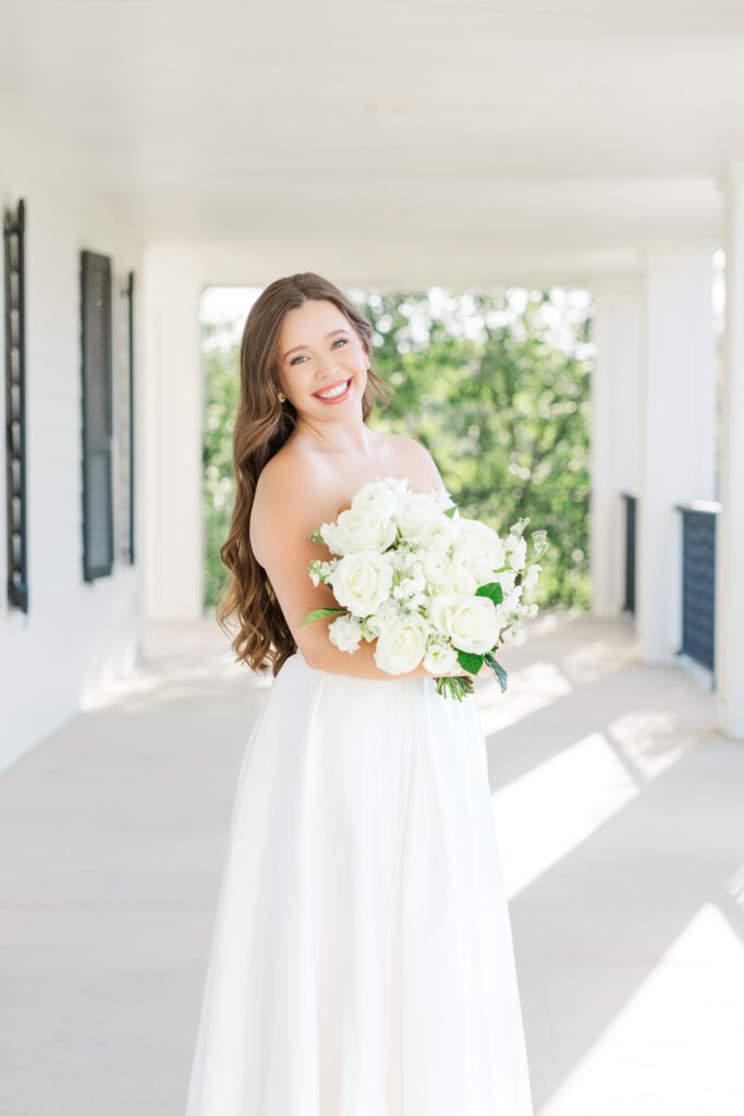 San Antonio bride smiles at the camera while holding a bouquet of white flowers on the porch at Kendall Point venue.