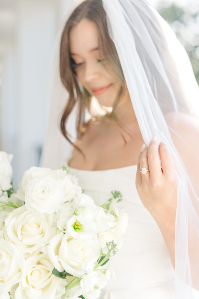 this is a bride who is holding her veil and showing off her ring finger with abeautiful opal ring and looking down at her white bouquet of flowers 