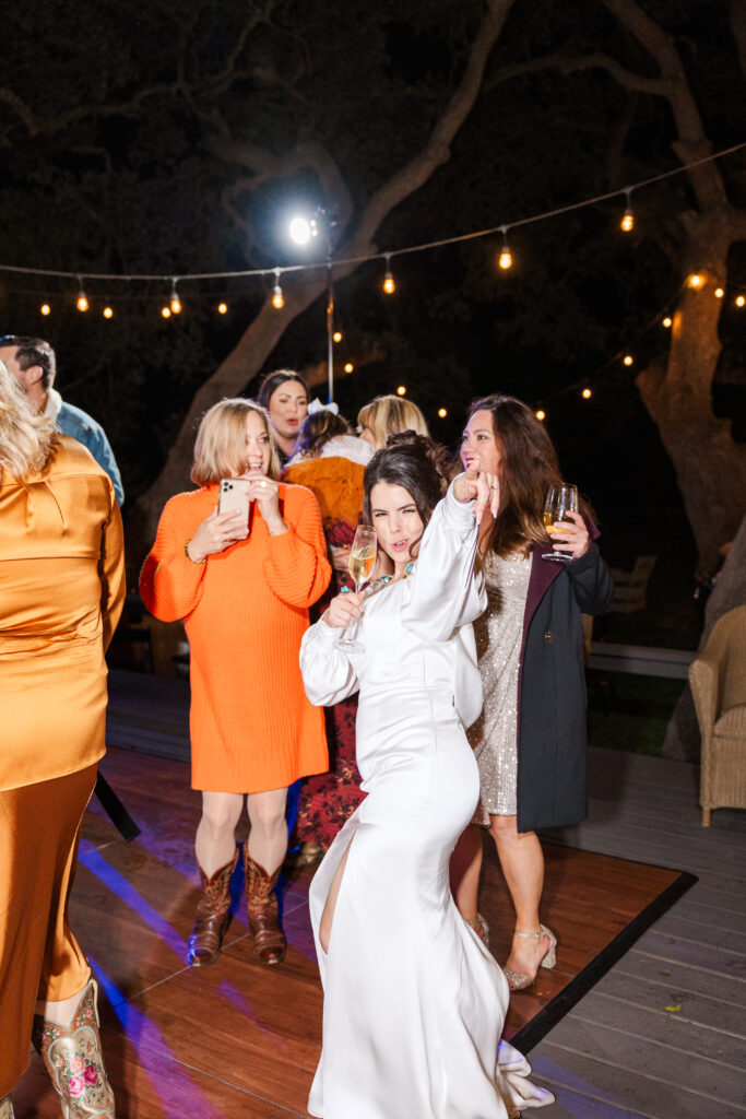 Bride poses for the camera with friends in orange dresses dancing behind her. It's almost the wedding night