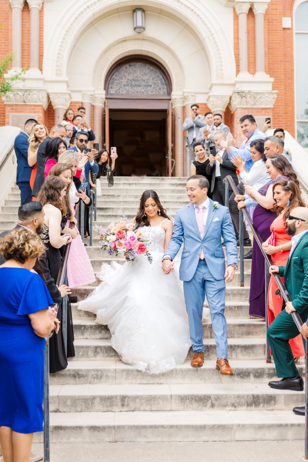 Crowd looks on as bride holds bouquet with one hand and grooms hand in the other as they walk down a grand stair case leading out of the San Antonio wedding venue