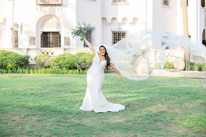 Bride holds bouquet up in the air and has the other arm up as her veil blows in the wind. She shows off makeup done by a wedding make up artist in San Antonio. The background is a castle like building.