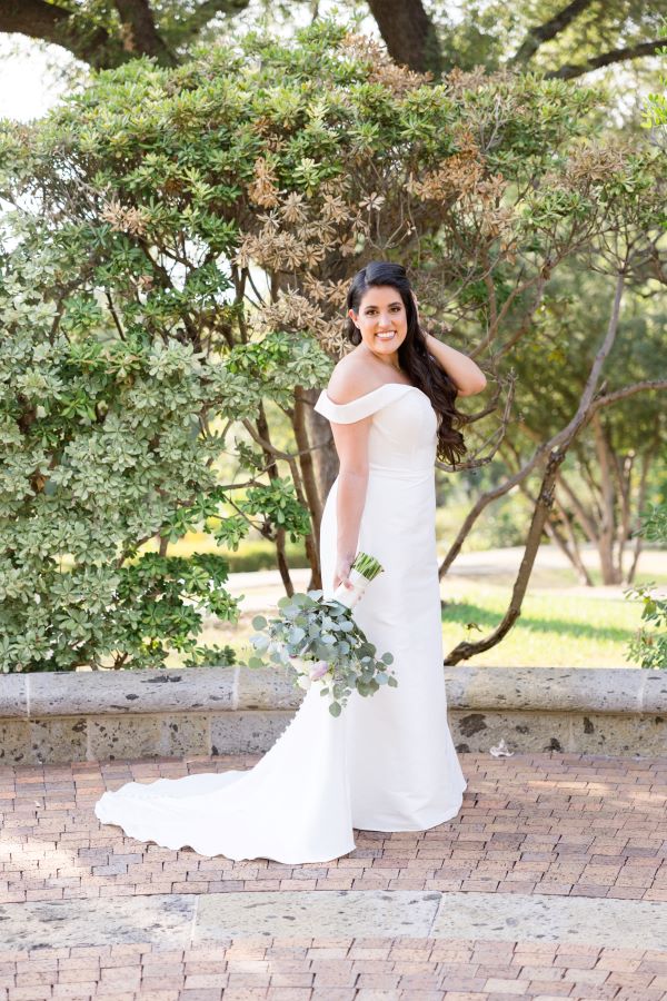 San Antonio wedding makeup artist does an incredible job doing a bride's makeup. In the picture the bride looks confident, playing with her hair and standing up against some trees.