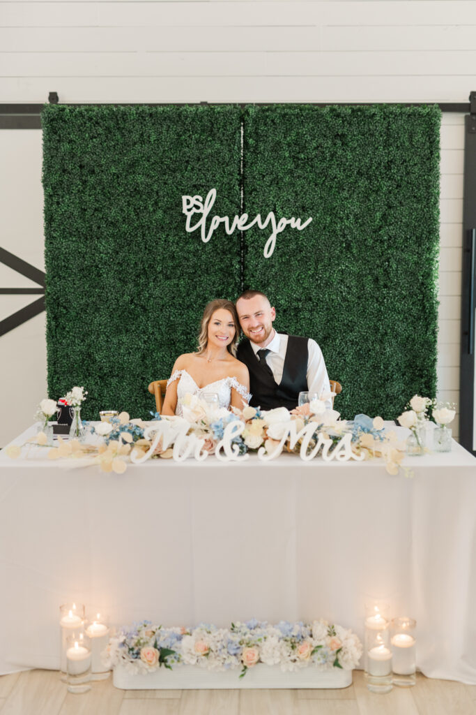 Dallas bride and groom sit at their sweetheart table with the words "p.s I love you" behind them at The Gardenia venue