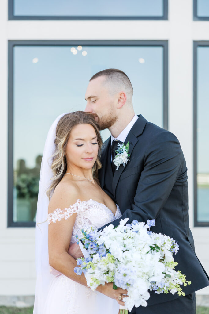 Dallas groom kisses the Dallas bride's forehead as she holds a white and blue flower bouquet right outside The Gardenia Venue