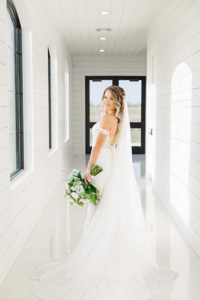 Blonde bride stands in an all white hallway with her back facing the camera, head turned over her shoulder to look into the camera. She is in a white form-fitting wedding dress with a cathedral style veil and a bouquet of white and blue flowers.