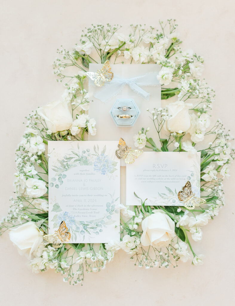 Gold plastic butterflies sit on the wedding invitations with a ring box with an engagement ring and wedding band toward the top of the photo lay on top of white flowers for San Antonio wedding flat lay