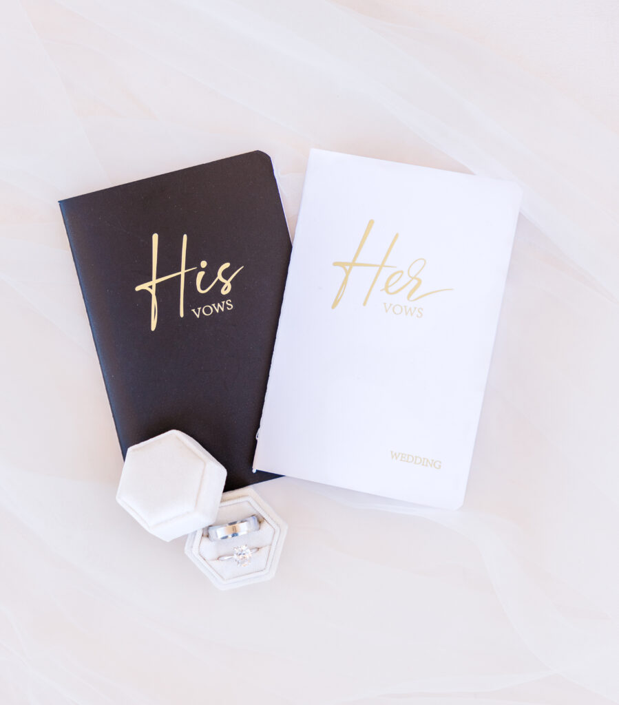 His and Hers vow books and a ring box with a wedding band and an engagement ring lay on a mat for a wedding flat lay photo